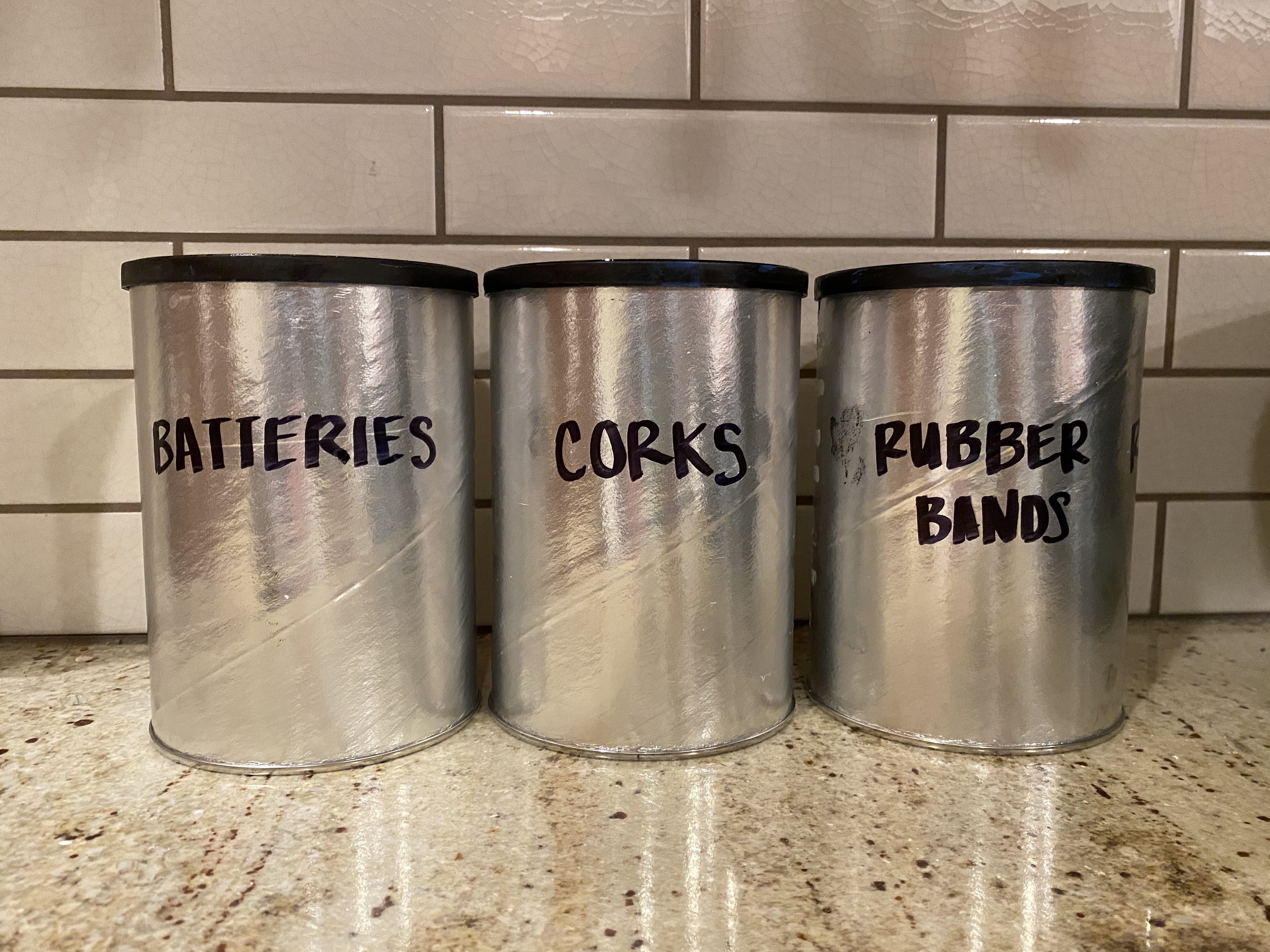 three silver cyclinders labeled rubberbands, batteries, and corks