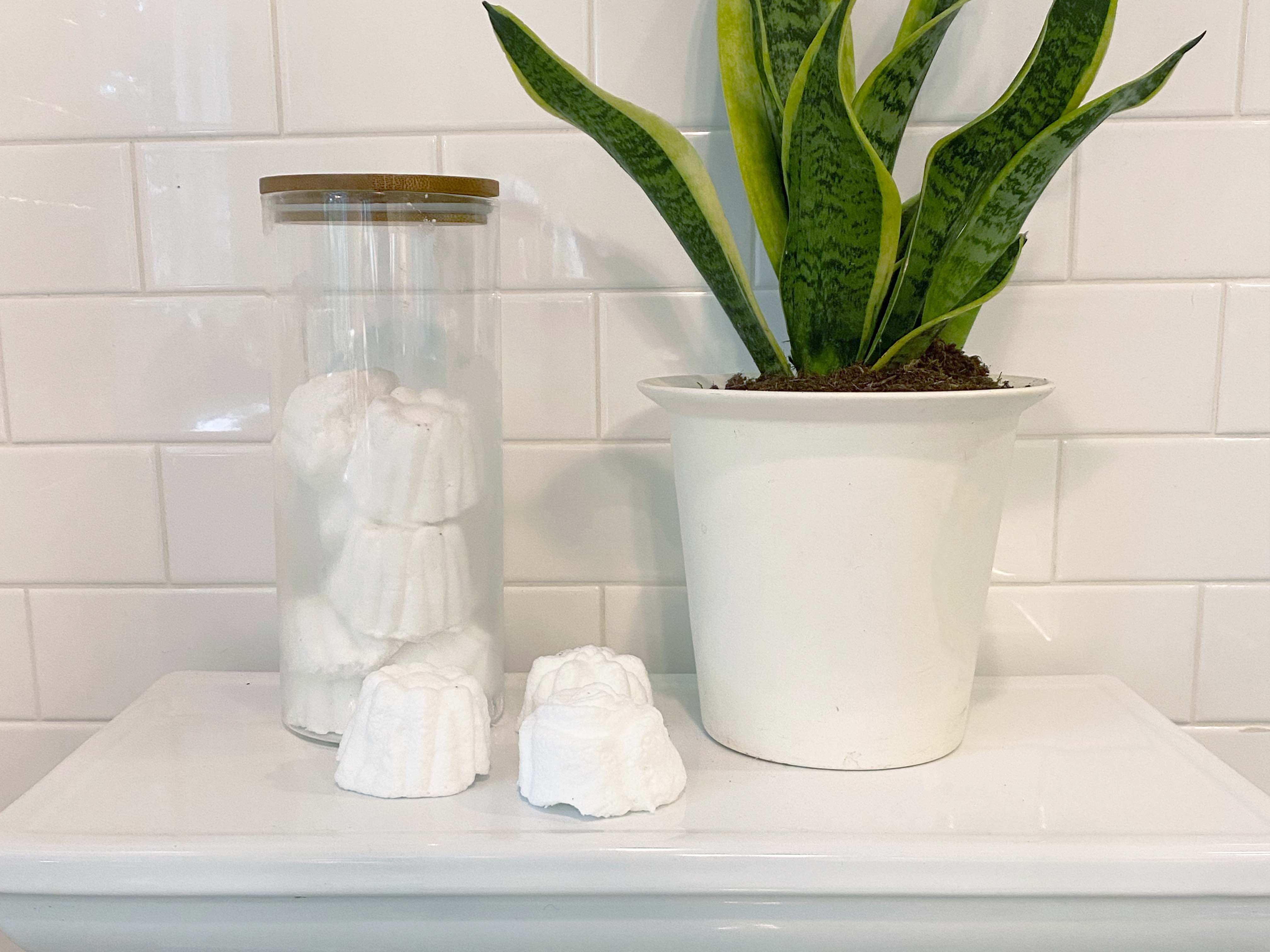 three toilet bombs next to glass jar full of toilet bombs and house plant all on top of toilet tank