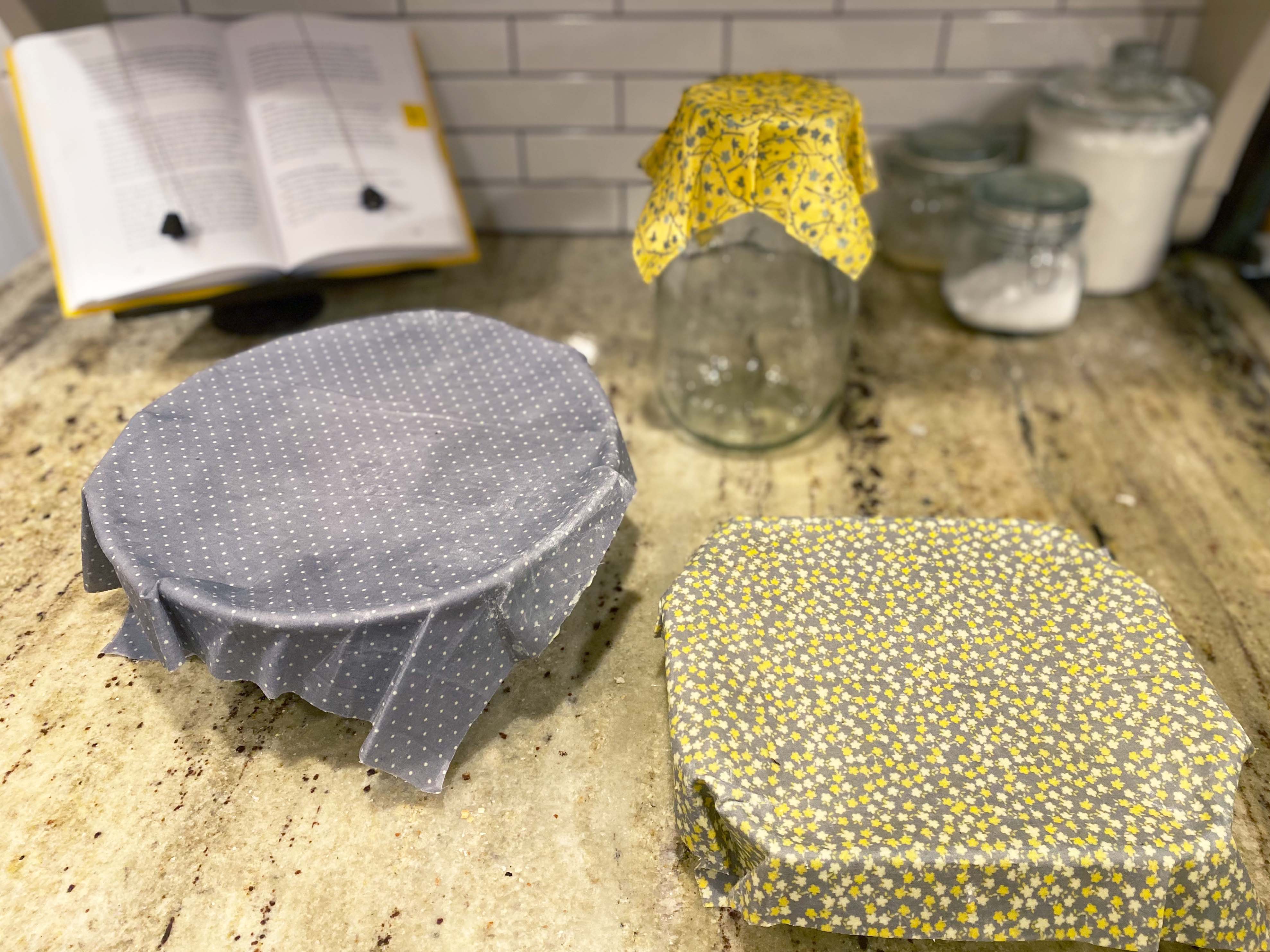 glass jar, bowl, and casserole dish on counter with homemade beeswax wraps over tops. cookbook and flour jar blurry in background